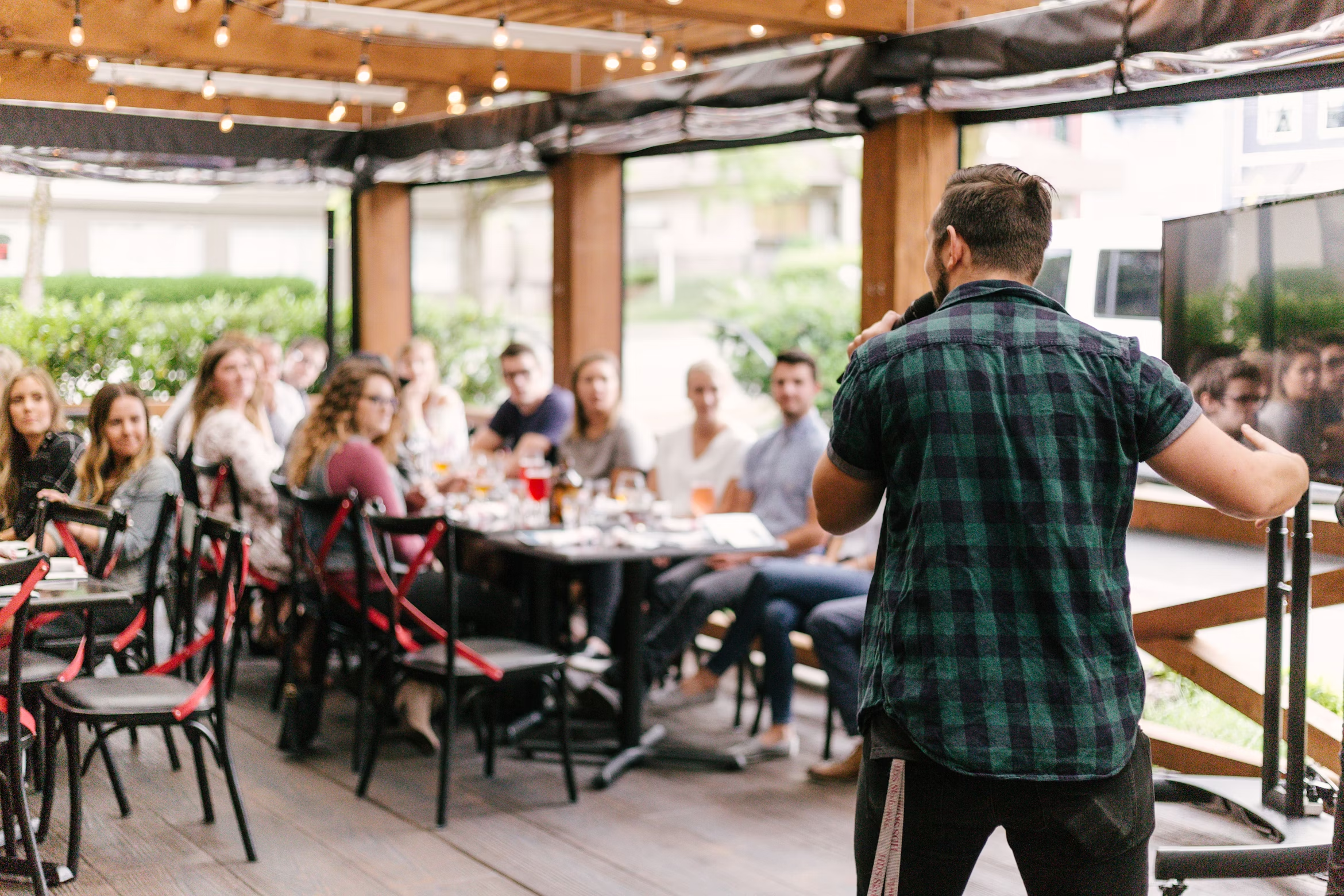 A person speaks to a seated group at an outdoor gathering area, with string lights overhead and people listening around tables,
